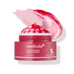 Un pot rose ouvert étiqueté "Medicube Crème Capsule TXA Niacinamide 55 g" révèle des capsules roses et blanches à l'intérieur, avec un frottis rose-blanc crémeux et une capsule translucide montrée au-dessus du pot. Dakar