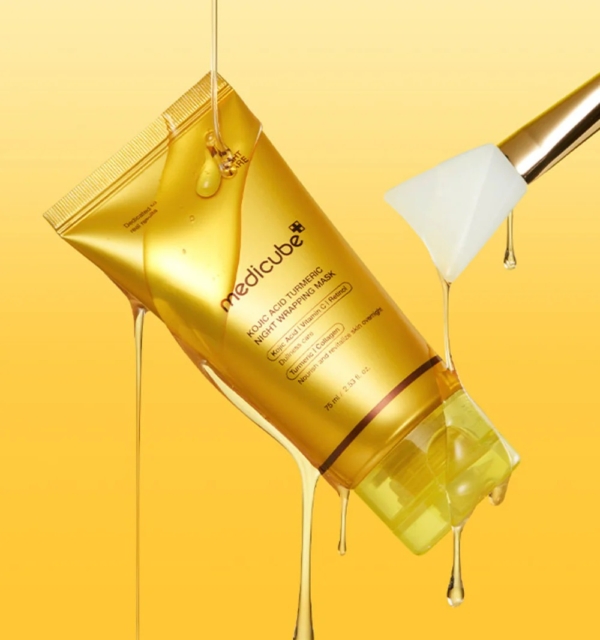 Un tube doré de Medicube Masque de Nuit Révélateur d'Éclat Acide Kojique et Curcuma 75 ml se trouve au milieu de gouttes de miel sur un fond jaune, avec une spatule munie d'un masque blanc placée à droite. Dakar