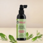 Un flacon noir de Mielle Organics Huile nettoyante fortifiante cuir chevelu (Romarin Menthe) 118ml se tient debout à côté de WAAM Paris Huile de Moringa, entouré de feuilles fraîches de romarin et de menthe sur un fond beige clair. Dakar