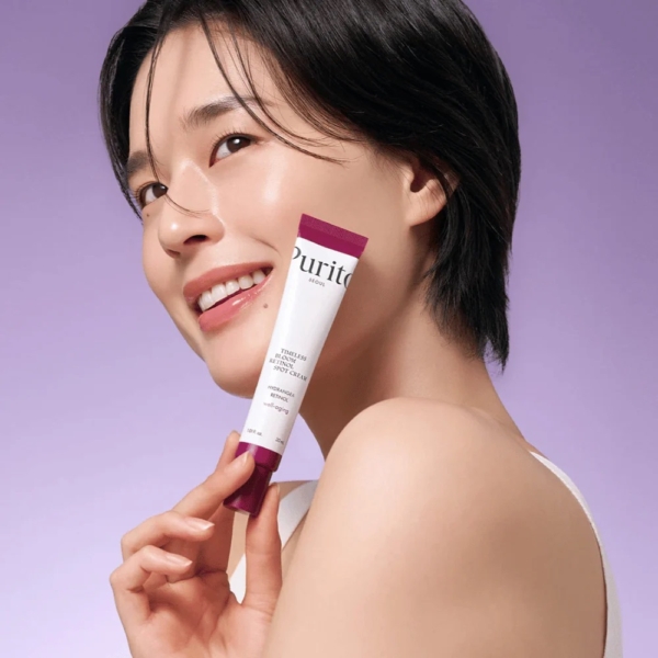 Une personne aux cheveux courts et bruns sourit, tenant Purito Crème Anti Taches au Rétinol 0,1% Timeless Bloom 30 ml près de son visage sur un fond violet clair. Elle porte un haut blanc et a une peau claire et rayonnante. Dakar