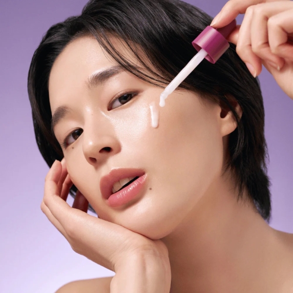 Une personne aux cheveux bruns courts et au maquillage naturel applique Purito Sérum Lissant Anti Age 1% de Bakuchiol 30 ml sur sa joue à l'aide d'un compte-gouttes. Sa peau apparaît lisse et rosée sur un fond dégradé violet. Dakar