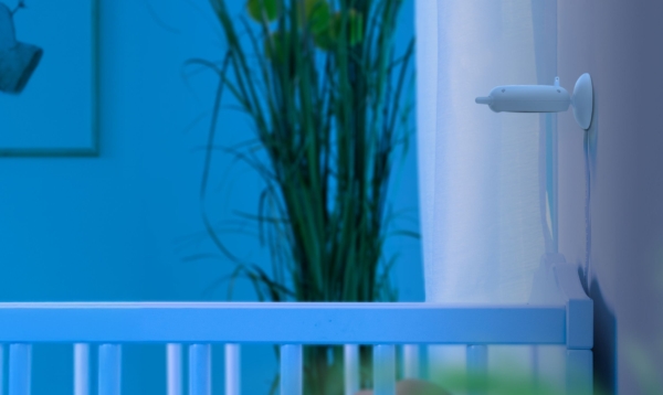 La Reer BabyCam S Moniteur Vidéo pour Bébé est installée sur un mur bleu clair au-dessus d'un berceau, à côté d'une grande plante verte et d'un rideau transparent dans une chambre d'enfant aux tons bleus et à l'éclairage tamisé. Dakar
