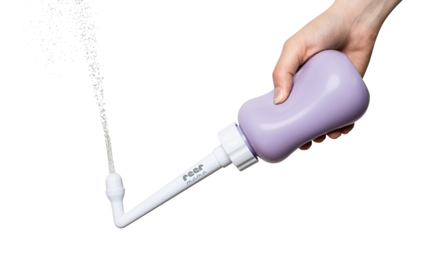 Une main tient le Reer Douche Intime Sensible Maman, un flacon compressible violet avec un embout blanc incliné, pulvérisant un fin filet d'eau, sur un fond blanc. Dakar