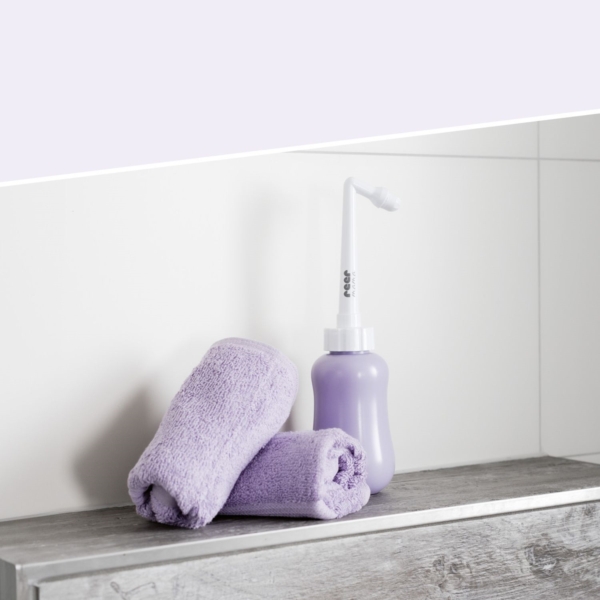 Une Reer Douche Intime Sensible Maman est posée à côté de deux serviettes de lavande soigneusement enroulées sur une étagère de salle de bains en bois, contre un mur carrelé blanc. Dakar