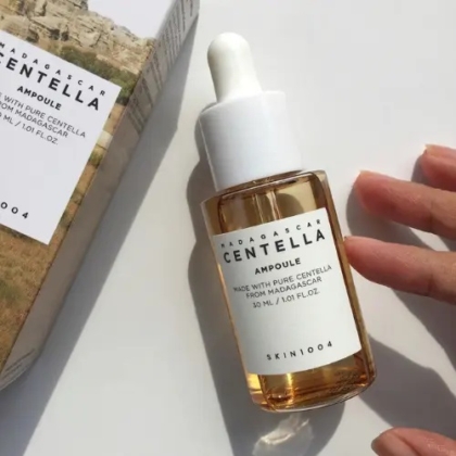 Une main se tend vers un flacon compte-gouttes transparent étiqueté "Skin1004 Madagascar Centella Ampoule Sérum Apaisant et Anti Imperfections 30 ml", contenant un liquide jaune clair, reposant sur une surface blanche à côté de sa boîte. Dakar
