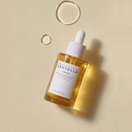 Un flacon de Skin1004 Madagascar Centella Ampoule Sérum Apaisant et Anti Imperfections 30 ml avec un bouchon compte-gouttes blanc repose sur une surface beige à côté de trois gouttes de liquide, projetant une ombre douce. Dakar