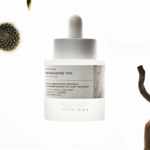 Un flacon compte-gouttes givré étiqueté "Skin1004 Sérum Éclaircissant Intensif à la Niacinamide et aux Micro Spicules 30" se tient debout sur un fond blanc brillant, flanqué de formes végétales abstraites partiellement visibles. Dakar