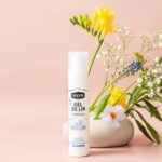 Un flacon de WAAM Gel de Lin BIO 100ml est présenté devant un vase blanc rempli de fleurs jaunes et blanches, le tout sur un fond rose tendre. Dakar