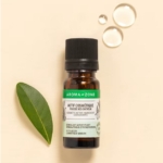 Un petit flacon foncé d'Aroma Zone Actif Pousse des Cheveux 10ml repose sur une surface beige avec une feuille verte et deux gouttes à proximité, évoquant un cosmétique naturel pour la pousse des cheveux. Dakar