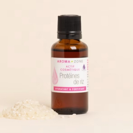 Un flacon en verre brun d'Aroma Zone Actif Protéines de Riz hydrolysées 30ml avec un bouchon noir se trouve à côté d'un petit tas de riz non cuit sur un fond beige clair. Dakar