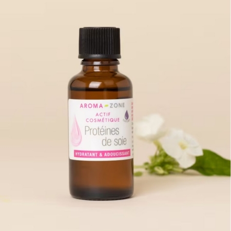 Un flacon en verre brun d'Aroma Zone Actif Protéines de Soie hydrolysées 5ml avec un bouchon noir repose sur une surface beige près d'une fleur blanche et de feuilles vertes, destiné à un usage cosmétique et hydratant. Dakar