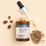 Flacon ambré d'huile à barbe Aroma-Zone à l'acide hyaluronique et au chanvre biologique, un compte-gouttes, des graines de chanvre et plusieurs noix ou graines brunes disposées sur un fond beige clair. Dakar