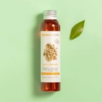Une bouteille d'Aroma Zone Huile de Fenugrec BIO 100ML se trouve sur un fond vert clair à côté d'une feuille verte. Le flacon transparent révèle une huile ambrée, et son étiquette blanche présente des images de graines de fenugrec. Dakar