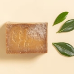 Aroma Zone Savon d'Alep 25% Laurier 200g, un savon naturel brun avec des bulles en surface, est présenté à côté de trois feuilles vertes sur un fond beige. Dakar