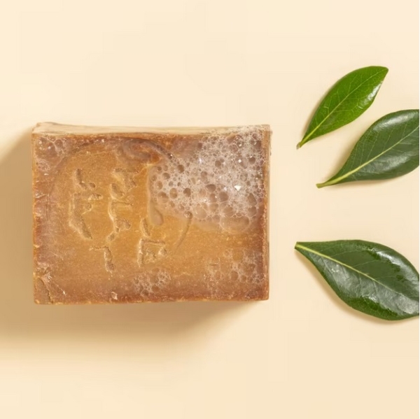 Aroma Zone Savon d'Alep 25% Laurier 200g, un savon naturel brun avec des bulles en surface, est présenté à côté de trois feuilles vertes sur un fond beige. Dakar
