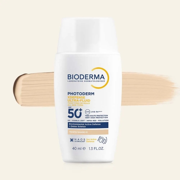 Le Fluide Solaire Teinté Photoderm XDefense SPF50 Ultra Fluide 40ml de Bioderma est présenté à l'endroit devant un produit beige sur un fond clair, avec des étiquettes mettant l'accent sur la protection de l'environnement et de la pollution. Dakar