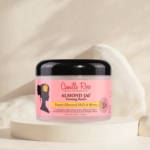 Le pot de Camille Rose Almond Jai Crème Cheveux pour les Boucles 240 ml se trouve sur un fond neutre, avec une étiquette rose au texte noir et la silhouette d'une femme avec une fleur dans les cheveux. Dakar