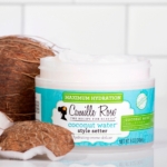 Un pot ouvert de Crème Coiffante Hydratante Deluxe à l'Eau de Coco 240 ml de Camille Rose présente sa formule crémeuse blanche, avec une noix de coco coupée en deux et des morceaux de noix de coco disposés sur une surface carrelée blanche à proximité. Dakar