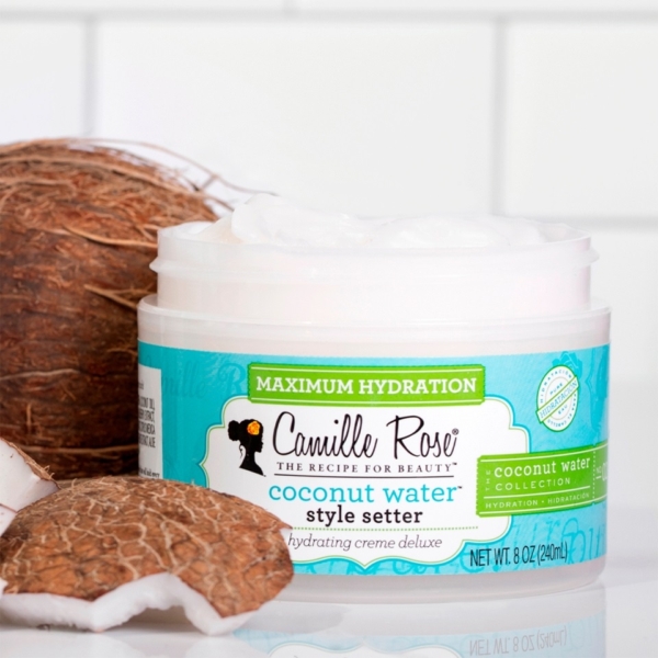 Un pot ouvert de Crème Coiffante Hydratante Deluxe à l'Eau de Coco 240 ml de Camille Rose présente sa formule crémeuse blanche, avec une noix de coco coupée en deux et des morceaux de noix de coco disposés sur une surface carrelée blanche à proximité. Dakar
