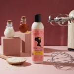 Un flacon de 240 ml de Camille Rose Curl Love Lait Hydratant est disposé sur une surface rose avec un fouet, du riz, un mixeur, des petits pots de lait et de miel, et un bol d'huile à côté de blocs pastel. Dakar