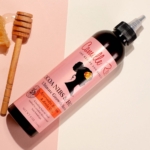 Un flacon de Camille Rose Sérum fortifiant COCOA NIBS et HONEY 240ml repose sur une surface claire à côté d'une cuillère à miel en bois. L'étiquette rose présente une silhouette de femme et un motif floral. Dakar