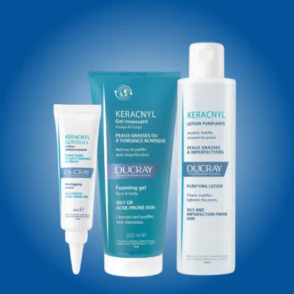 Trois produits de soin Ducray Keracnyl pour les peaux grasses et à imperfections sont présentés sur un fond bleu dégradé : un tube, un gel moussant et un flacon de lotion purifiante, disponibles chez Univers Cosmetix. Dakar