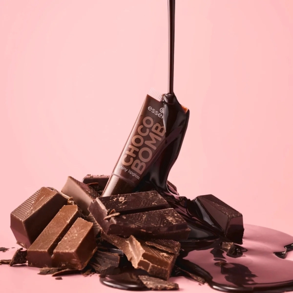 Un tube de Essence Gloss à Lèvres Choco Bomb 01 Chocoholic 10 ml se trouve sur un fond rose, entouré de morceaux de chocolat et recouvert d'un épais filet de chocolat fondu. Dakar