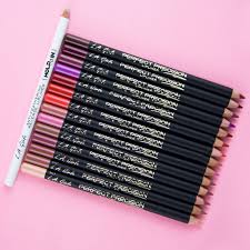 Quatorze crayons L.A. Girl Perfect Precision LipLiner en noir et un en blanc, chacun de 1,49 g, sont disposés en diagonale sur un fond rose, montrant une gamme de nuances allant du nude au rouge et au violet. Dakar