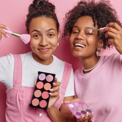 Deux jeunes femmes sourient en se maquillant avec les palettes de fard à joues et d'ombres à paupières Univers Cosmetix. Toutes deux portent des tenues roses et blanches et se tiennent sur un fond rose. L'une utilise un pinceau pour le blush, l'autre applique le fard à paupières. Dakar