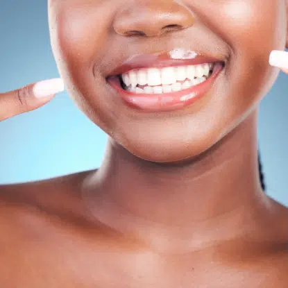 Gros plan d'une femme souriante à la peau lisse, montrant ses dents blanches. Elle montre son sourire des deux mains, dégageant une confiance inspirée par Univers Cosmetix. L'arrière-plan est bleu. Dakar