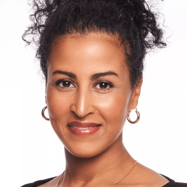Une femme aux cheveux noirs bouclés attachés, portant des boucles d'oreilles et un haut noir, sourit doucement à l'appareil photo sur un fond blanc, son regard radieux étant rehaussé par la Crème Soleil Fondant Bronzeur Teint Hâlé Fini Mat 9 g de Catrice. Dakar