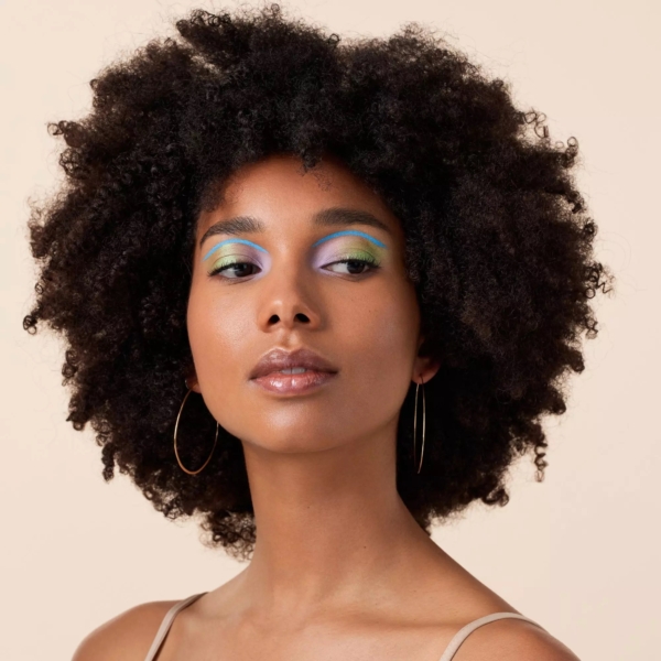 Une femme aux cheveux bouclés naturels, avec de grandes boucles d'oreilles et un maquillage des yeux bleu pastel, vert et violet, rayonne avec la crème solaire Catrice Melted Sun Crème Bronzeur Teint Hâlé Fini Mat 9 g alors qu'elle regarde sur le côté, sur un fond beige neutre. Dakar