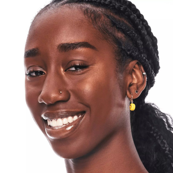 Une femme aux cheveux tressés, avec un piercing dans le nez et plusieurs boucles d'oreilles - dont une boucle en argent, une étoile et un pendentif jaune - affiche un sourire radieux. Son regard radieux suggère qu'elle a utilisé la palette Essence Illuminateurs Teint Rayonnant Pure Nude Sunlighter 40 Be My Sunlight. Dakar