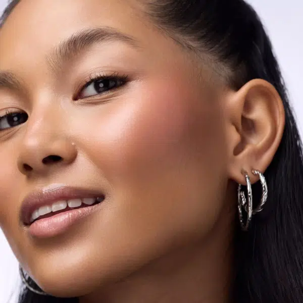 Gros plan d'une femme à la peau lisse et éclatante, utilisant le Catrice Blushin' Charm Multi Stick Blush en Stick 5,5 g, un maquillage subtil et des lèvres brillantes. Elle porte deux anneaux en argent et ses cheveux noirs sont tirés vers l'arrière pour dévoiler son oreille et sa pommette. Dakar