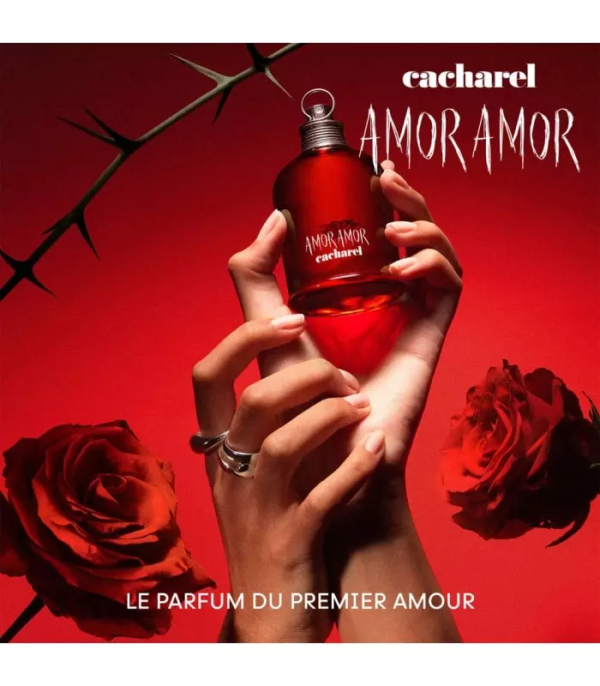 Une main tient un flacon rouge de Cacharel Amor Amor Eau de Toilette 30ml sur un fond rouge avec des roses et une branche épineuse. Le texte blanc indique "cacharel AMOR AMOR" et "LE PARFUM DU PREMIER AMOUR". Dakar