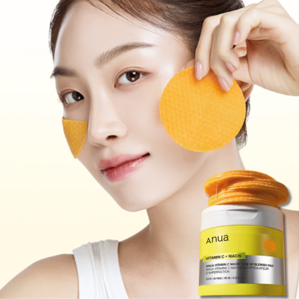 Une femme tient un tampon facial orange sur sa joue, tandis qu'un autre repose sur l'autre. Devant elle, un pot étiqueté "Anua Pads Éclaircissant et Exfoliant à la Vitamine C et Niacinamide 60 pads" contient d'autres coussinets orange. Dakar