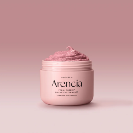Un pot rose clair étiqueté "Arencia Fresh Rosehip Rice Mochi Soin 3 en 1 Nettoyant Exfoliant et Masque 120 g", rempli d'un nettoyant fouetté rose, sur fond de dégradé rose lisse. Dakar