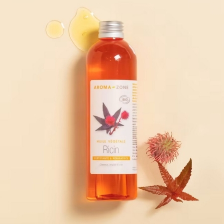 Un flacon d'Aroma Zone Huile de Ricin 250 ml repose sur une surface beige clair, avec à proximité de l'huile renversée, une feuille de ricin et une cosse de graine. Dakar