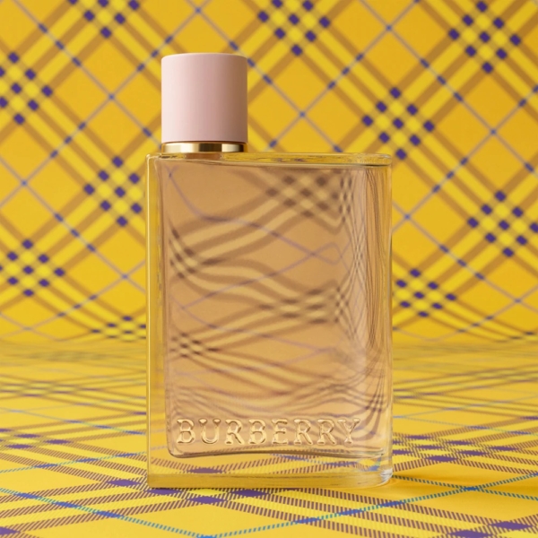 Un flacon transparent de Burberry Her Eau de Parfum pour Femme 50ml avec un bouchon rose pâle repose sur une surface à carreaux jaunes, sur un fond assorti. Le logo Burberry est embossé sur le devant du flacon. Dakar