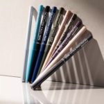 Neuf crayons Catrice 20H Ultra Precision Gel Waterproof Crayon Yeux Gel 0,08 g, chacun dans une teinte métallique ou mate unique, sont disposés en une rangée ordonnée sur une surface légère et réfléchissante avec des ombres douces. Dakar