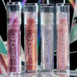 Quatre tubes de Catrice Diamond Glaze Lip Gloss Brillant 3 ml se tiennent debout devant des surfaces prismatiques, présentant des nuances chatoyantes de rose, de cuivre, d'argent irisé et de pêche claire. Dakar