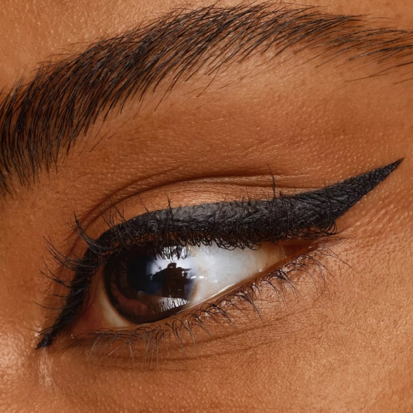 Gros plan sur un œil à la peau brune et lisse, aux cils épais et à l'aile noire dramatique et nette, réalisé avec l'Eyeliner 24h Brush Liner Noir Longue Tenue 3 ml de Catrice ; le sourcil complet est soigneusement entretenu. Dakar