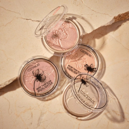 Trois poudriers ouverts Catrice More Than Glow Highlighter Poudre Compacte Illuminateur 5,9 g de différentes teintes sont présentés sur une surface beige craquelée, chacun présentant un motif floral gaufré sur la poudre. Dakar