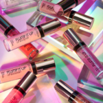 Plusieurs tubes de Catrice Plump It Up Lip Booster Gloss Effet Repulpant 3,05 ml sont présentés sur une surface vibrante et irisée avec des reflets colorés et des formes géométriques, créant une scène glamour. Dakar