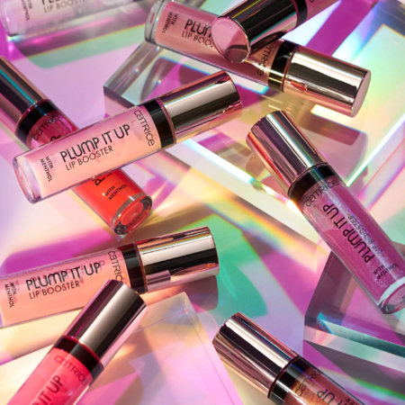 Plusieurs tubes de Catrice Plump It Up Lip Booster Gloss Effet Repulpant 3,05 ml sont présentés sur une surface vibrante et irisée avec des reflets colorés et des formes géométriques, créant une scène glamour. Dakar
