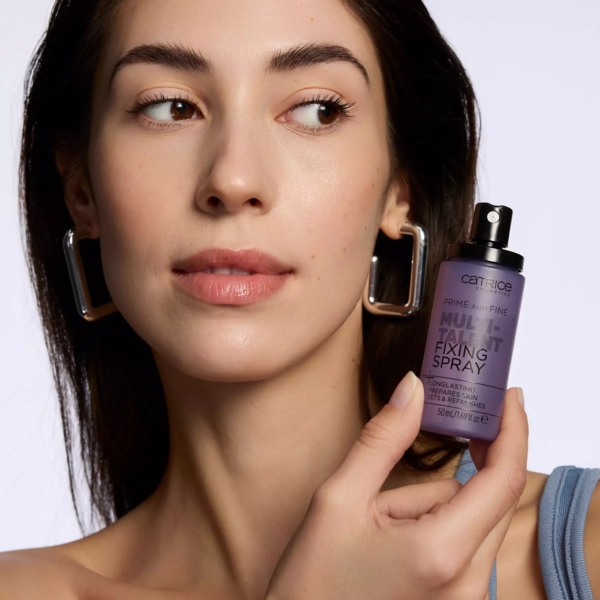 Une femme à la peau claire et aux grandes boucles d'oreilles carrées tient un flacon violet de Catrice Spray Fixateur Maquillage Multifonction Prime And Fine Multitalent Fixing 50 ml près de son visage, en regardant légèrement sur le côté. Dakar