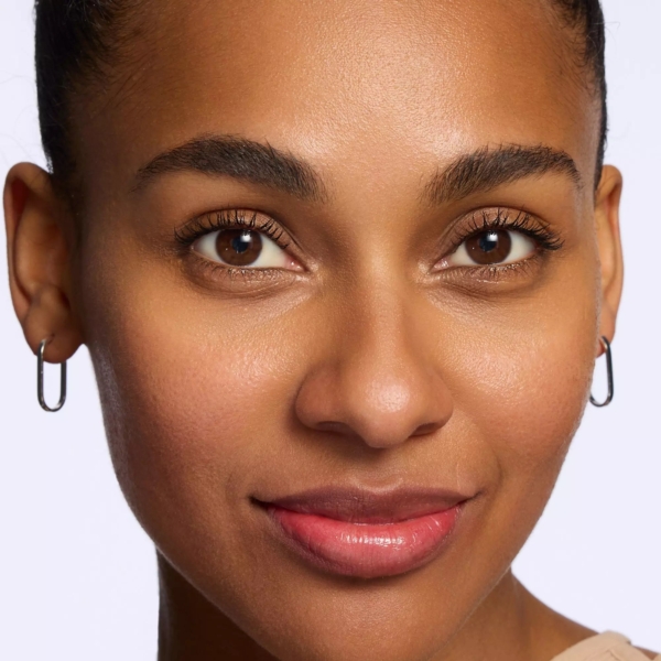 Un gros plan d'une femme à la peau lisse et éclatante et aux lèvres brillantes montre son look impeccable obtenu avec le Spray Fixateur Maquillage Multifonction Prime And Fine Multitalent Fixing 50 ml de Catrice. Elle a les yeux bruns, des sourcils bien dessinés, de petits anneaux et un fond clair uni. Dakar