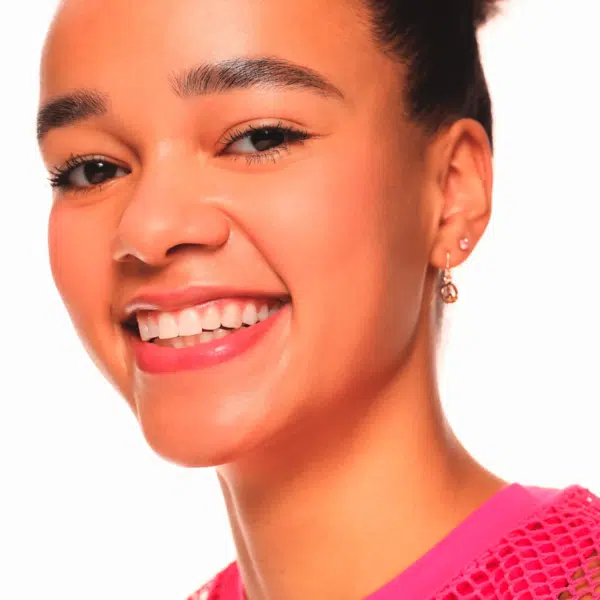Une jeune femme au teint moyen sourit vivement en montrant ses dents. Elle porte un haut rose et des boucles d'oreilles dorées. Ses cheveux sont tirés en arrière et l'arrière-plan est blanc. Dakar