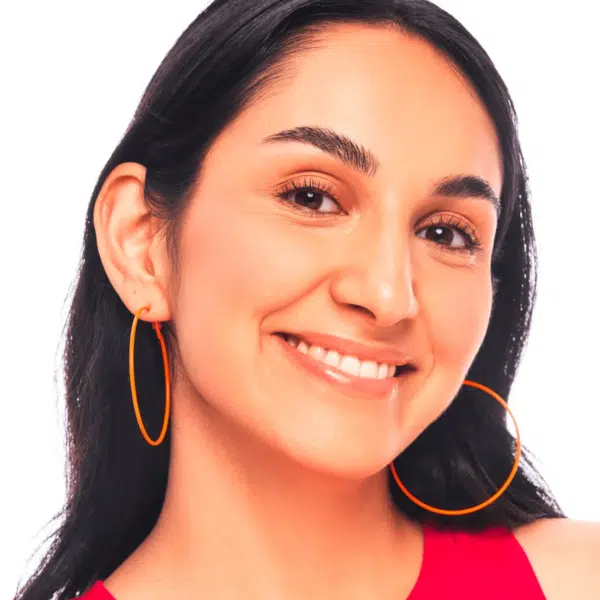 Une femme aux longs cheveux bruns sourit, portant Essence Blush Fini Mat Love Is In The Air 10 Dream Up 8g, de grandes boucles d'oreilles orange et un haut rouge sans manches sur fond blanc. Dakar