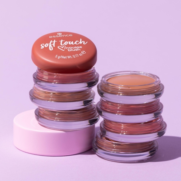 Deux pots en verre de blush mousse Essence Soft Touch aux côtés ondulés sont empilés sur une surface violette claire, l'un fermé par un couvercle rouge et l'autre ouvert, laissant apparaître un blush pêche à l'intérieur. Dakar